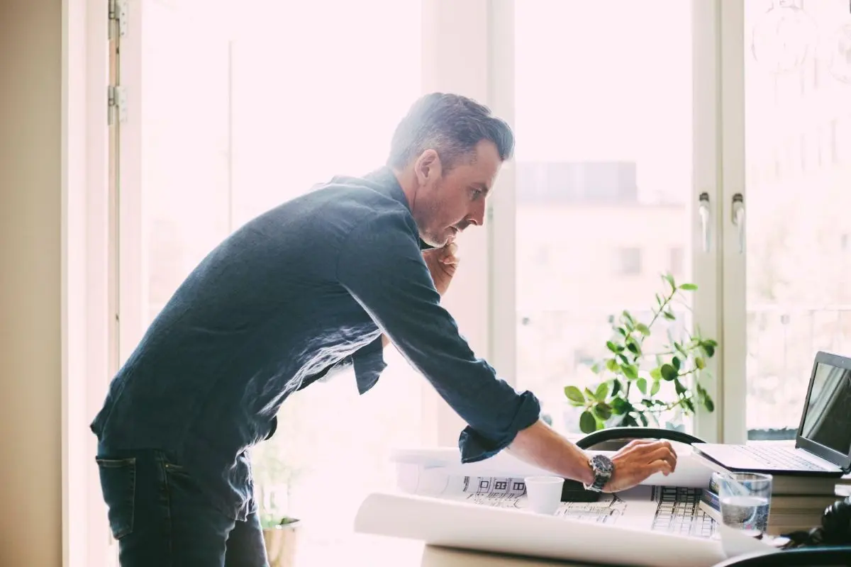 man standing over desk talking on phone and checking laptop screen